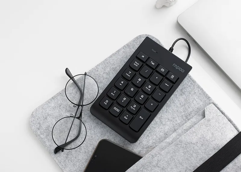 Rapoo K10 đặt trên túi nỉ xám cạnh kính mắt, điện thoại và laptop