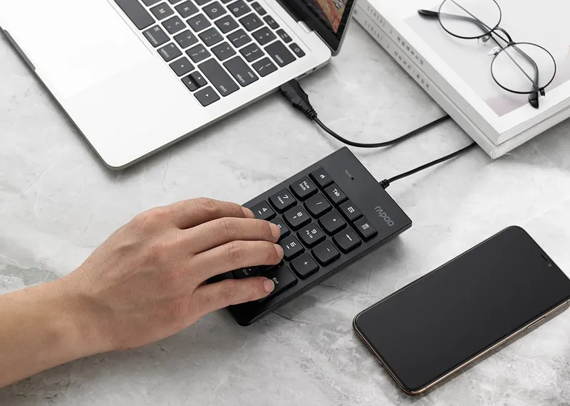 Tay đang gõ trên Rapoo K10 cắm dây vào laptop, bên cạnh là điện thoại